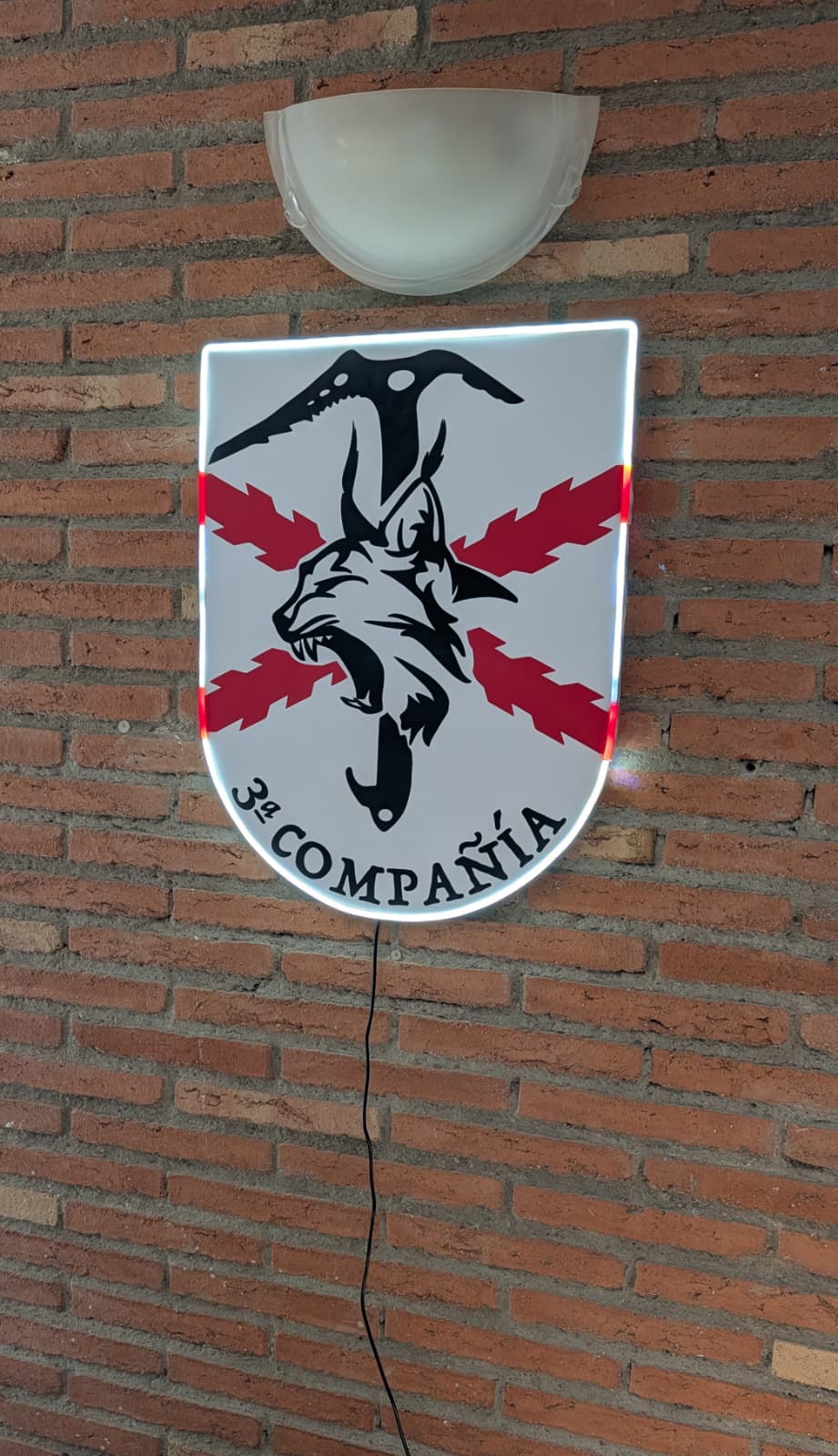 Cuadro decorativo con iluminación neón LED del escudo de la 3ª Compañía militar, montado sobre una pared de ladrillo visto. El diseño presenta un lince negro rugiendo sobre un piolet y la Cruz de Borgoña roja, con la inscripción "3ª COMPAÑÍA" en la base. El borde del escudo está realzado por una tira de neón blanco brillante que genera un efecto de retroiluminación.